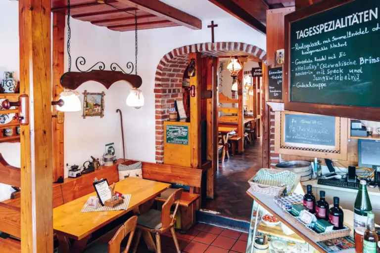 Gemütliches, rustikales deutsches Restaurant mit Holzbänken und -tischen, Hängelampen, einer Speisekarte auf Kreidetafeln und einer Auslage mit Flaschen und Gebäck in der Nähe des Eingangs.
