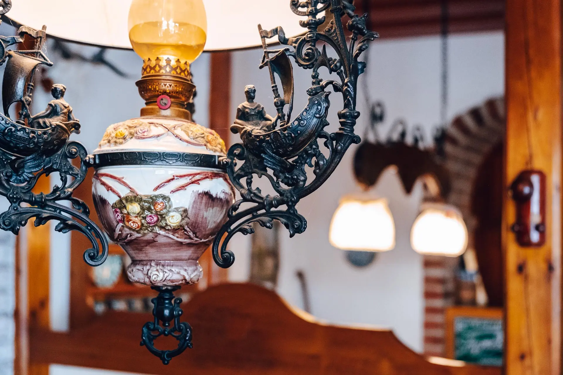 Nahaufnahme einer verschnörkelten Vintage-Lampe mit komplizierten Metallarbeiten und floralen Mustern, die in einem Raum mit verschwommenen Lichtern und Holzmöbeln im Hintergrund hängt.