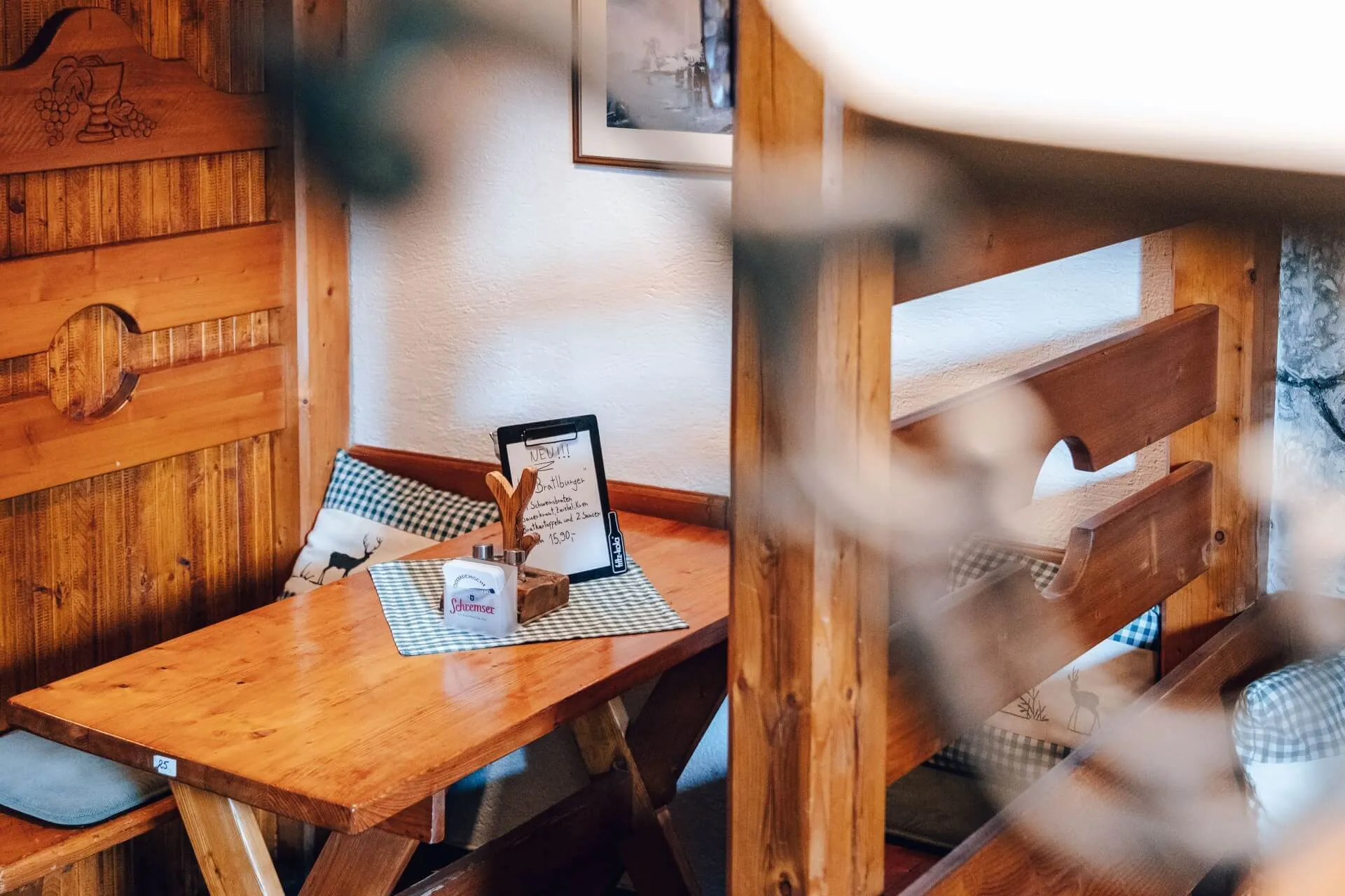 Ein gemütlicher Holztisch mit Bänken und karierten Kissen in einem rustikalen Café, dekoriert mit gerahmter Kunst und hölzernen Akzenten, sanft beleuchtet von natürlichem Licht.