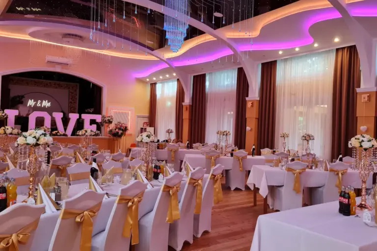 Großes Schutzhaus Rosental Ein eleganter Festsaal mit gedeckten Tischen für die Gäste, weißen Stühlen, die mit goldenen Schleifen verziert sind, Blumenschmuck auf den Tischen und einem großen, beleuchteten „LOVE“-Schriftzug unter sanfter violetter Beleuchtung.