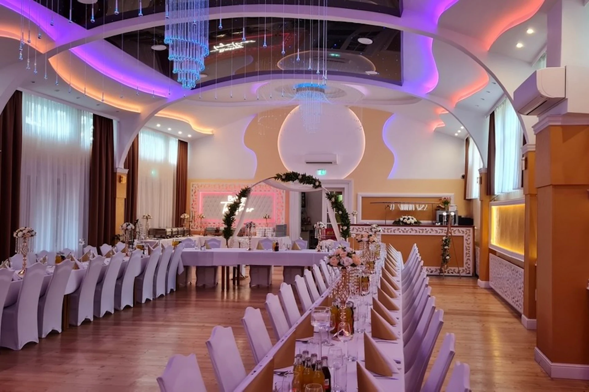 Ein eleganter Bankettsaal mit langen Tischen, die für eine Veranstaltung gedeckt sind, weißen Stühlen, Blumengestecken und bunten Deckenleuchten. Ein Bogen und Kronleuchter runden die Dekoration ab; im Hintergrund ist eine Bar zu sehen.