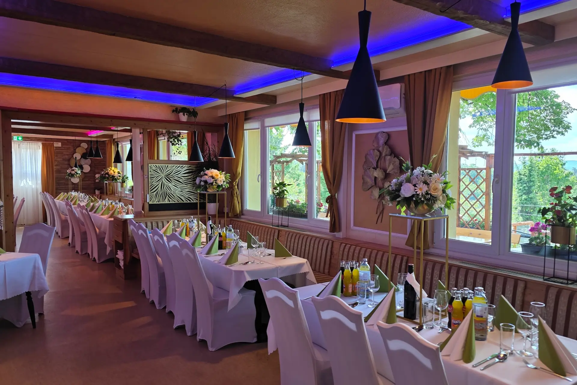 Ein eleganter Speisesaal mit langen Tischen, die für eine festliche Veranstaltung gedeckt sind; weiße Stühle, Blumengestecke, Getränke und violette Stimmungsbeleuchtung sorgen für eine festliche Atmosphäre an den großen Fenstern mit Blick ins Freie.