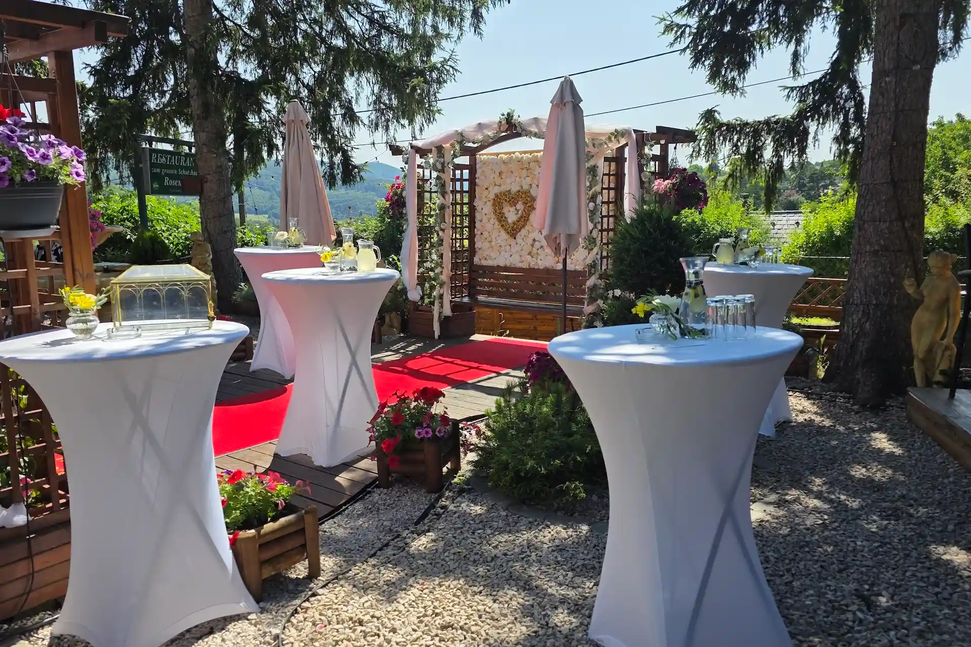 Hochzeitsdekoration im Freien mit Cocktailtischen, die mit weißen Tüchern gedeckt sind, Blumenschmuck und einem Holzbogen mit einer Hintergrundkulisse aus Herzspitzen. Ein roter Teppich führt zum Bogen, der von Grünpflanzen und Topfblumen umgeben ist.