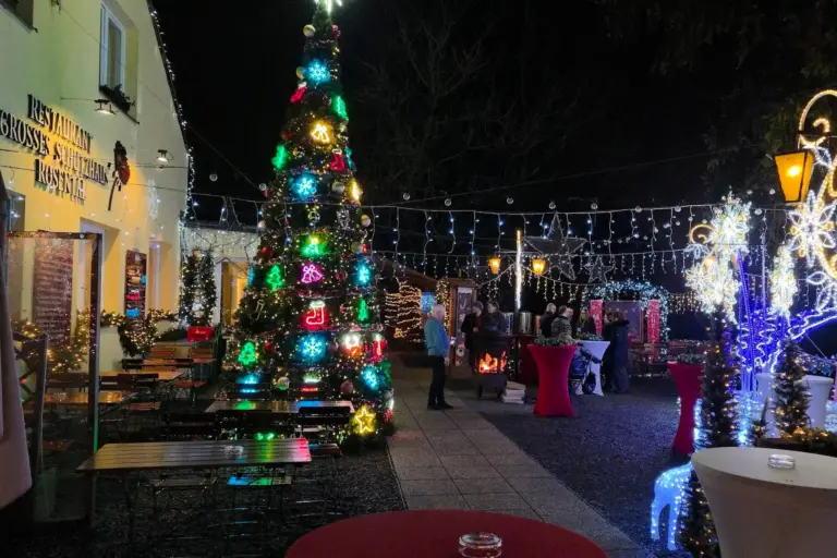 Eine festliche Szene im Freien bei Nacht mit einem geschmückten Weihnachtsbaum, Lichterketten, Tischen und Menschen, die sich um ein Lagerfeuer versammelt haben. Die Kulisse wirkt gemütlich und fröhlich, und die Weihnachtsdekoration taucht die Umgebung in ein festliches Licht.