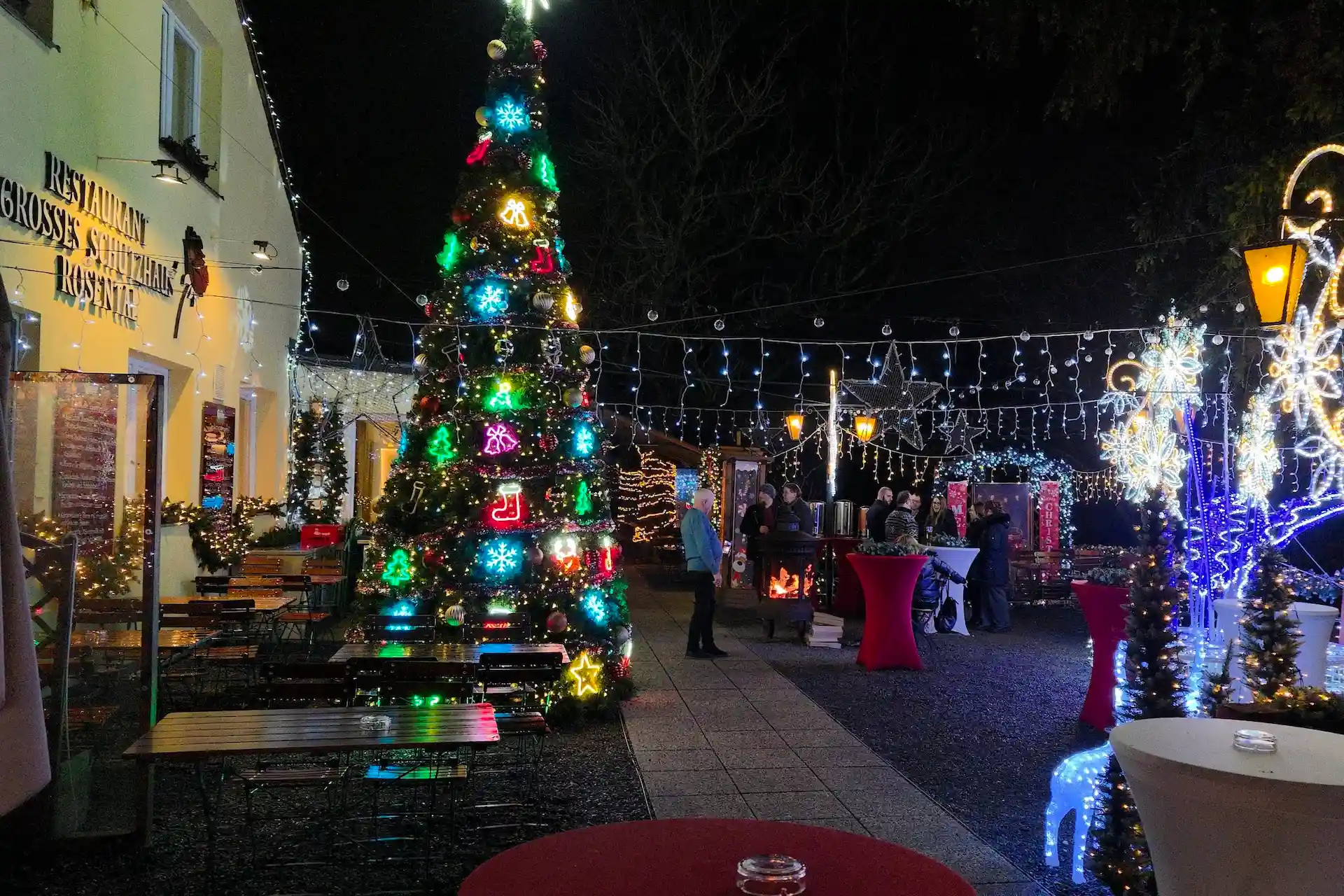Eine festliche Szene im Freien bei Nacht mit einem geschmückten Weihnachtsbaum, Lichterketten, Tischen und Menschen, die sich um ein Lagerfeuer versammelt haben. Die Kulisse wirkt gemütlich und fröhlich, und die Weihnachtsdekoration taucht die Umgebung in ein festliches Licht.