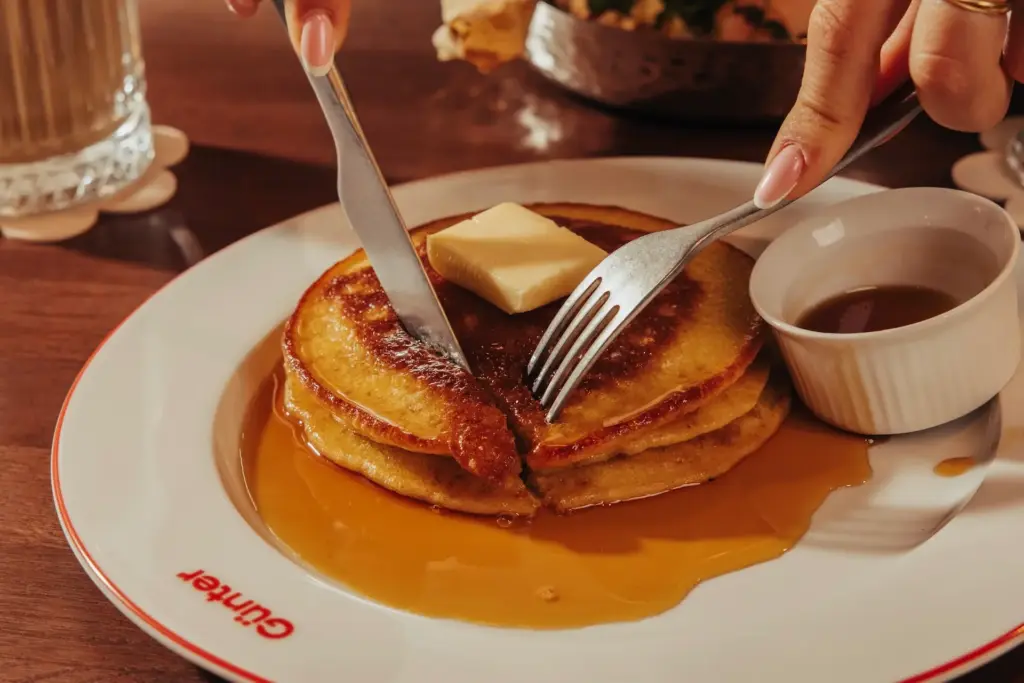 Eine Person schneidet mit Gabel und Messer in einen Stapel Pfannkuchen, die mit Butter bestrichen und mit Sirup beträufelt auf einem weißen Teller liegen, mit einer kleinen Schale Sirup an der Seite.