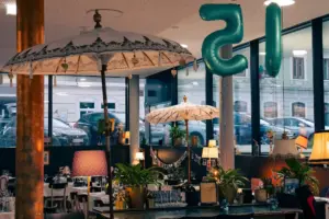 Das Habibi und Hawara, ein gemütlich eingerichtetes Café mit Vintage-Lampen, Topfpflanzen, weißen Sonnenschirmen und großen, blaugrünen Luftballons in Form der Zahl 15. Durch die großen Fenster sieht man draußen geparkte Autos und Gebäude.