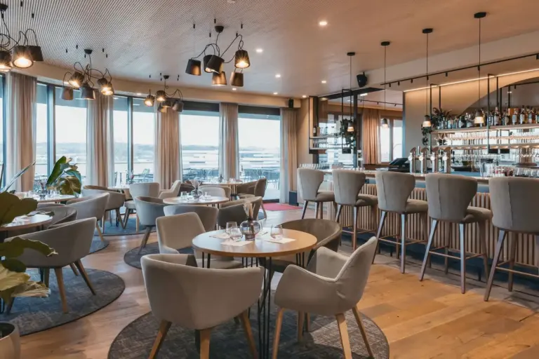 Hafnerbar Modernes Restaurantinterieur mit runden Tischen, gepolsterten Stühlen und einem Barbereich mit Barhockern, von dem aus man durch große Fenster den Ausblick genießen kann.