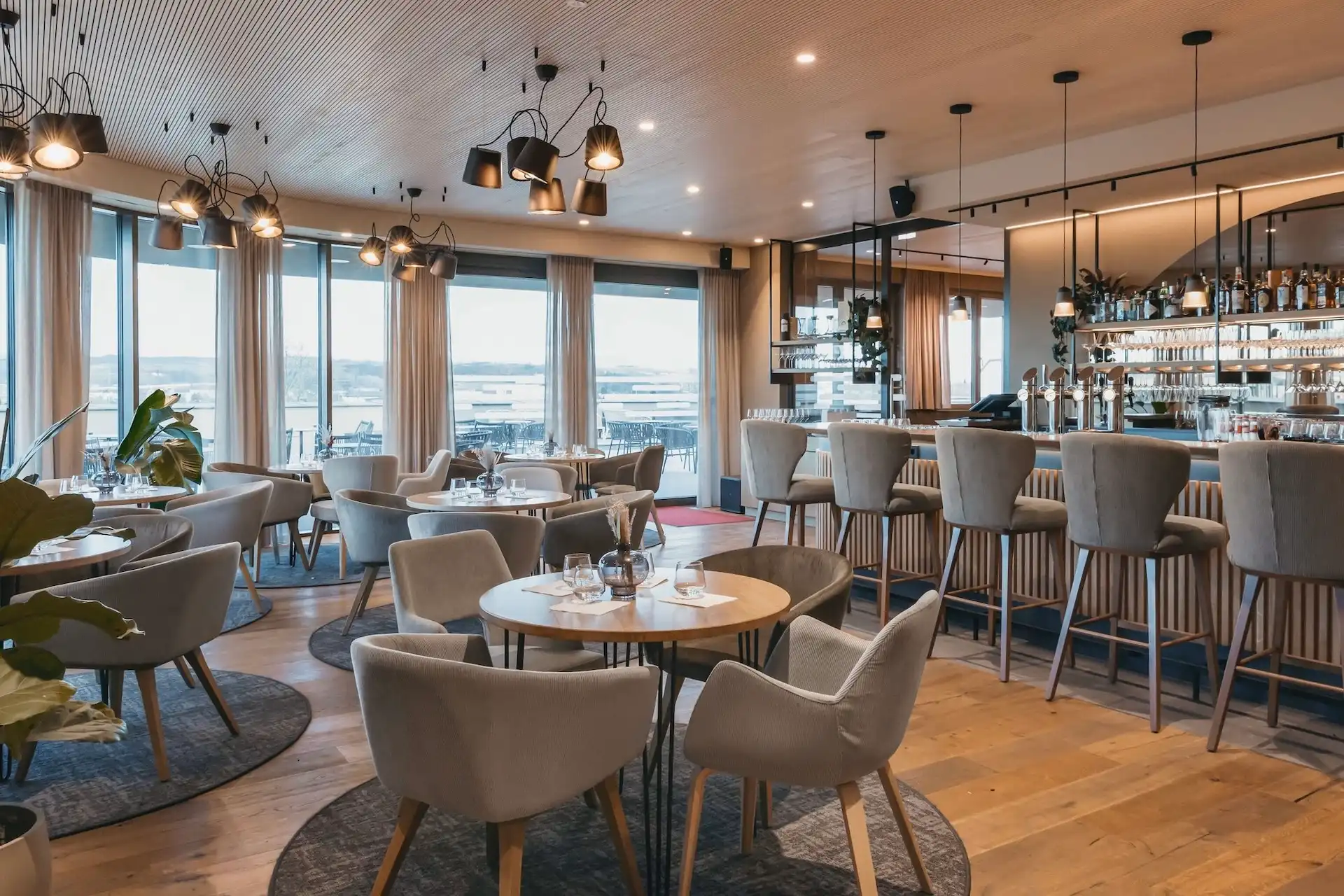 Hafnerbar Modernes Restaurantinterieur mit runden Tischen, gepolsterten Stühlen und einem Barbereich mit Barhockern, von dem aus man durch große Fenster den Ausblick genießen kann.