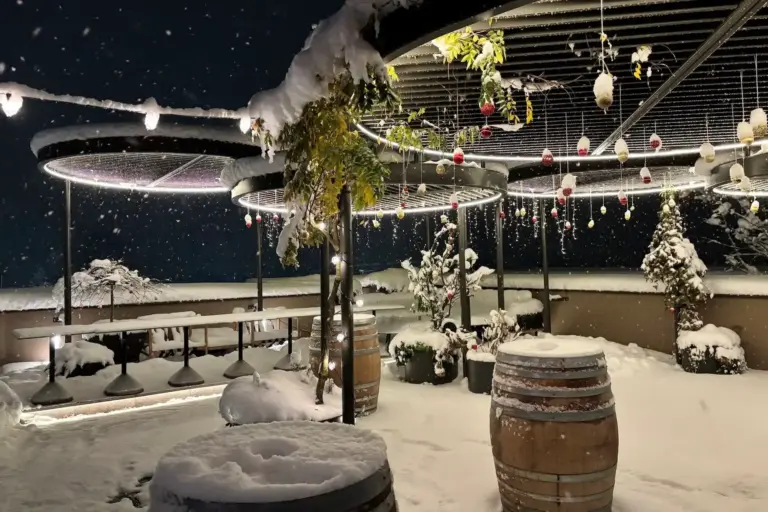 Eine schneebedeckte Terrasse bei Nacht, beleuchtet von runden Deckenleuchten und hängenden Dekorationen, mit Tischen aus Holzfässern.