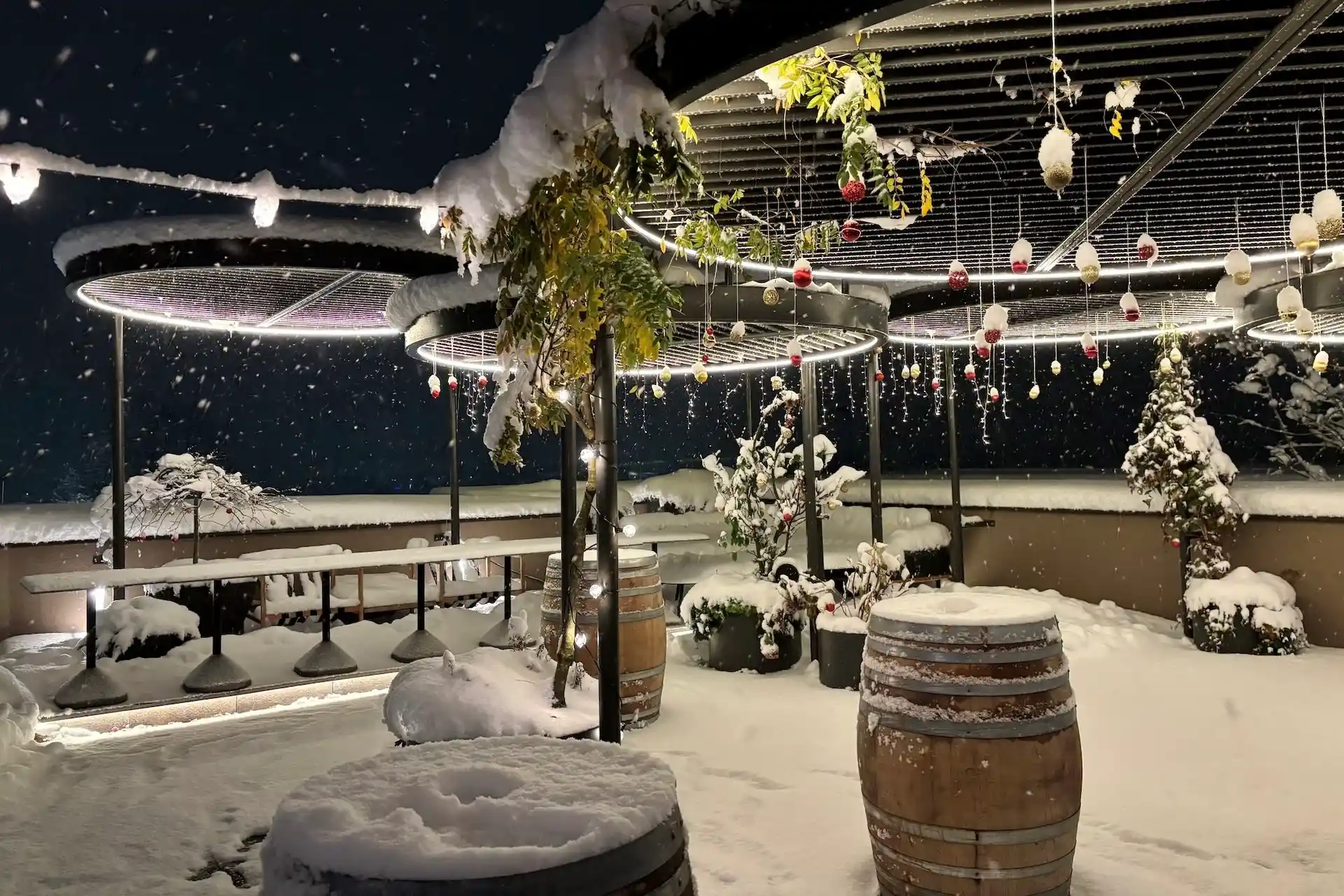 Eine schneebedeckte Terrasse bei Nacht, beleuchtet von runden Deckenleuchten und hängenden Dekorationen, mit Tischen aus Holzfässern.