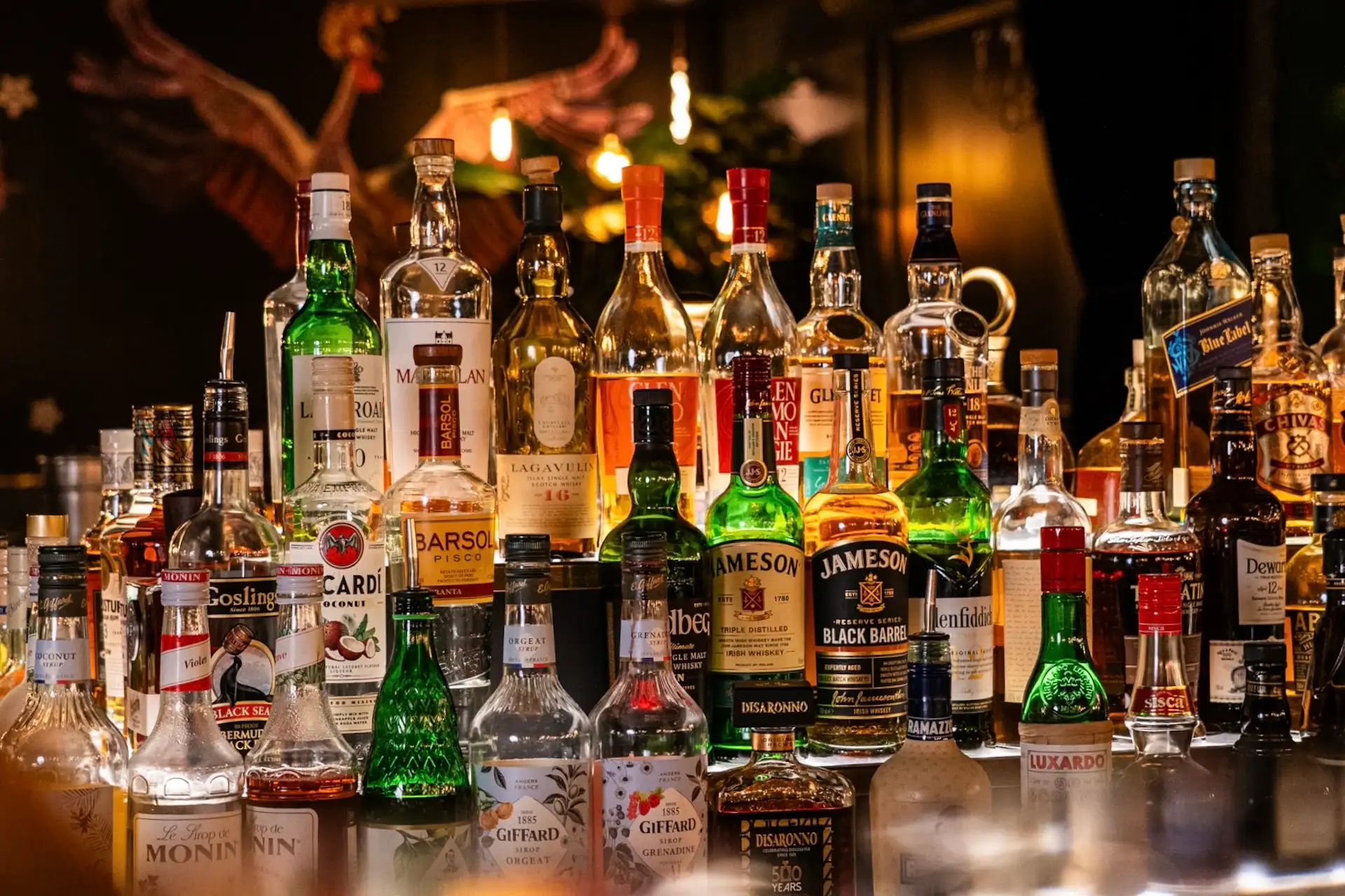 Verschiedene Spirituosenflaschen, darunter Whisky, Rum und Liköre, stehen dicht beieinander auf einer schwach beleuchteten Bartheke vor einem verschwommenen Hintergrund.