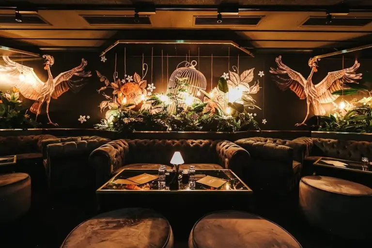 Eine luxuriöse Lounge mit dunklen Samtsofas, niedrigen Tischen und warmer, goldfarbener Beleuchtung. An der Wand hängen große Kunstwerke mit Kranichen und Blumenmotiven inmitten üppiger Grünpflanzen, die ein elegantes, einladendes Ambiente schaffen.