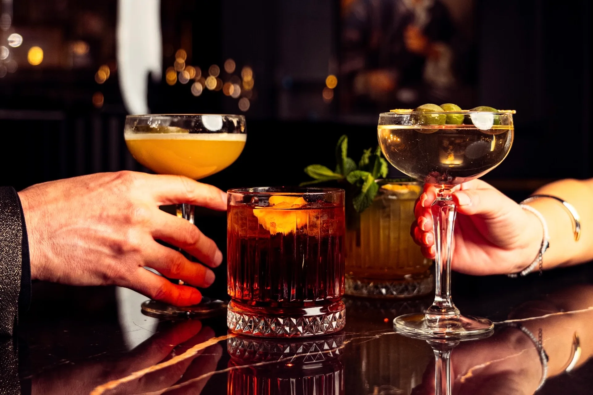 Zwei Personen, die elegante Cocktails in Kristallgläsern über einer spiegelnden, dunklen Bar halten, daneben weitere Cocktails und ein verschwommener, warm beleuchteter Hintergrund.
