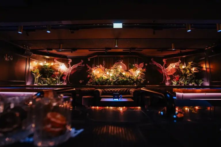 Eine schwach beleuchtete Lounge mit bequemen Sitzgelegenheiten, Zierpflanzen und einem Wandgemälde mit rosa Kranichen und Blumen auf schwarzer Wand, das eine luxuriöse, tropische Atmosphäre schafft. Auf der Bar im Vordergrund sind Gläser zu sehen.