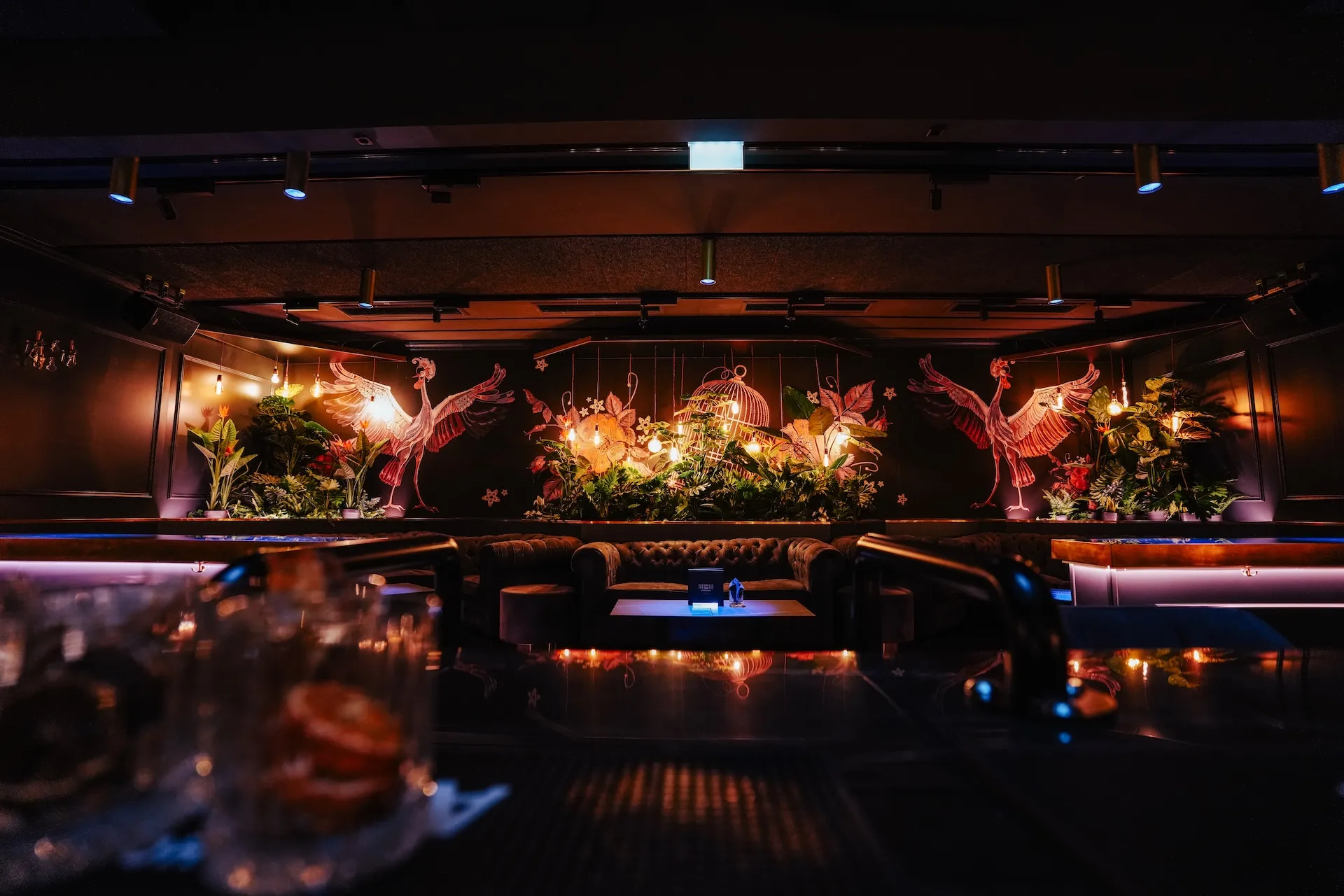 Eine schwach beleuchtete Lounge mit bequemen Sitzgelegenheiten, Zierpflanzen und einem Wandgemälde mit rosa Kranichen und Blumen auf schwarzer Wand, das eine luxuriöse, tropische Atmosphäre schafft. Auf der Bar im Vordergrund sind Gläser zu sehen.