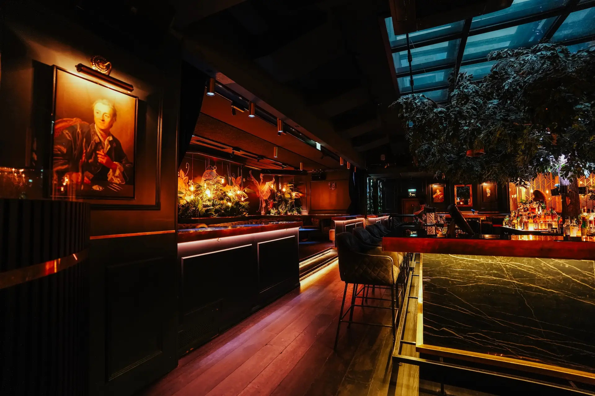 Eine schwach beleuchtete, gehobene Bar mit modernem Dekor, leuchtend orangefarbenen Lichtern, einer Marmortheke, Barhockern, einem Baum und gerahmten Kunstwerken an den dunklen Wänden, die eine gemütliche und elegante Atmosphäre schaffen.