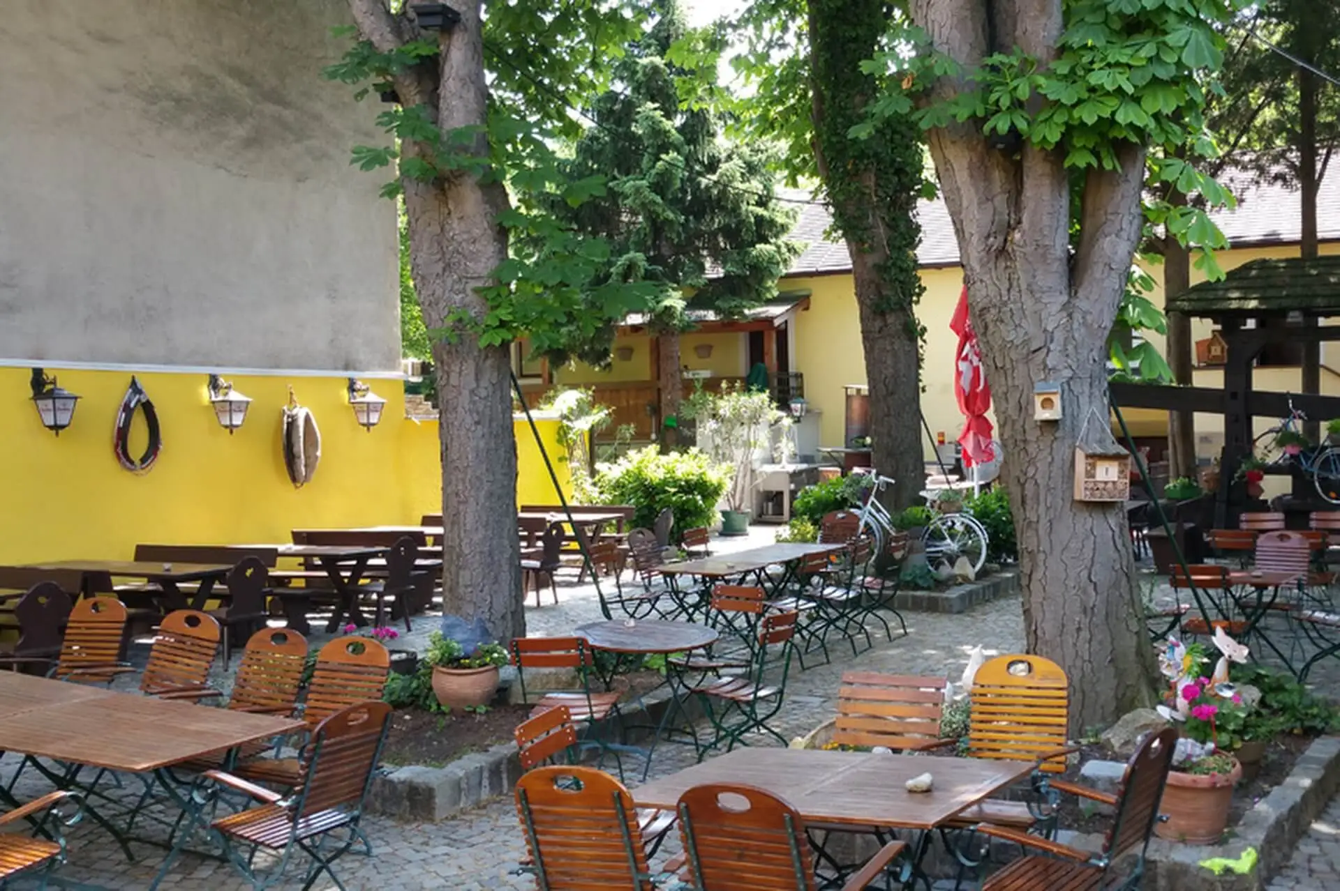 Außencafé mit leeren Holztischen und -stühlen auf einer Steinterrasse, die von großen Bäumen beschattet wird und von Grünflächen und gelben Gebäuden umgeben ist. Blumentöpfe und Dekorationen tragen zu einer gemütlichen, einladenden Atmosphäre bei.