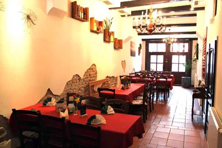 Ein gemütliches Restaurant mit roten Tischtüchern, Holzstühlen und rustikaler Einrichtung mit freiliegenden Ziegeln und Regalen mit Büchern und Pflanzen. Von der Decke hängen Kronleuchter, und durch die großen Fenster an der Vorderseite strömt natürliches Licht.