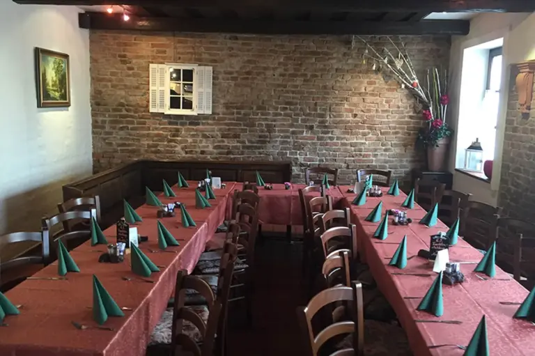 Ein gemütlicher Speisesaal mit Backsteinwänden und Holzstühlen um Tische mit roten Tischtüchern, auf jedem Platz liegen grüne gefaltete Servietten und kleine Dekorationen. Blumen und Pflanzen stehen in der Ecke neben einem Fenster.