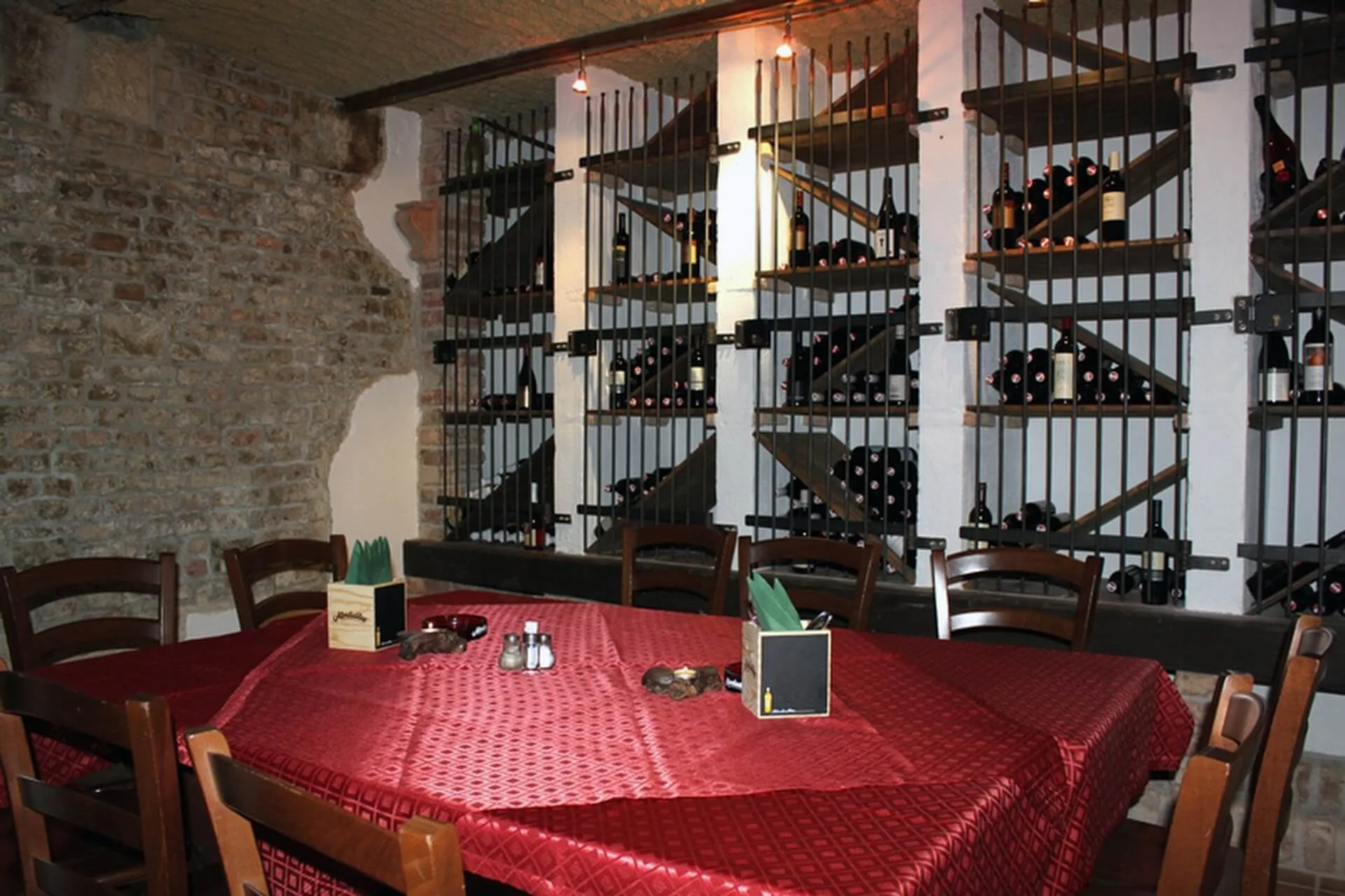 Ein gemütlicher Weinkeller mit freiliegenden Backsteinwänden, einem großen, mit roter Tischdecke bedeckten Tisch, Holzstühlen und Regalen voller Weinflaschen, die hinter Metallgittern gesichert sind. Die warme Deckenbeleuchtung trägt zur intimen Atmosphäre bei.