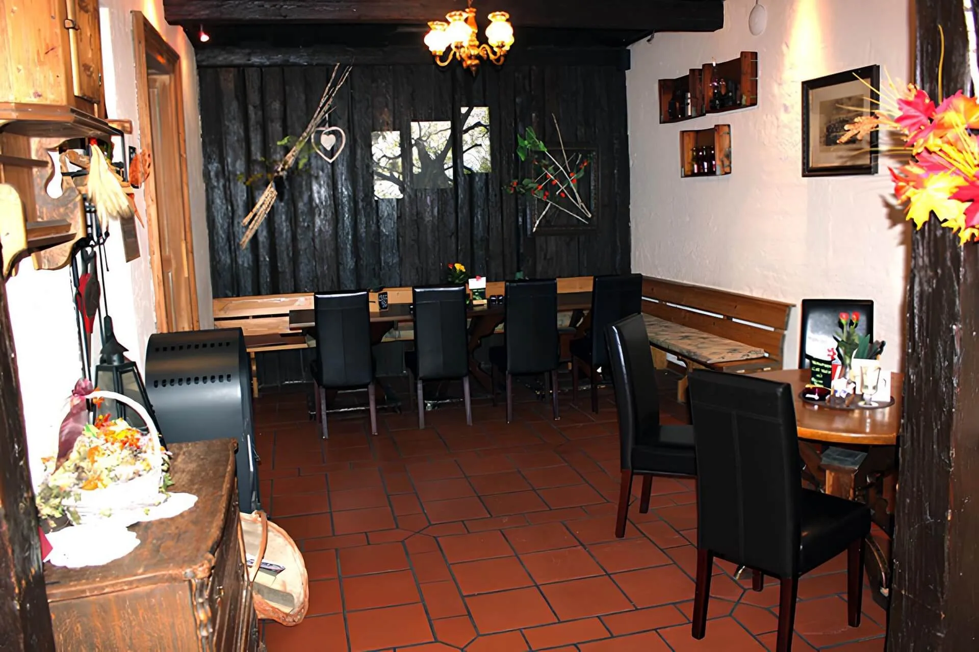 Ein gemütliches Restaurant mit schwarzen Holzwänden, schwarzen Stühlen an Holztischen, Herbstdekoration und einem Kronleuchter, der den Raum beleuchtet. An den Wänden befinden sich rustikale Regale und Dekorationsgegenstände.