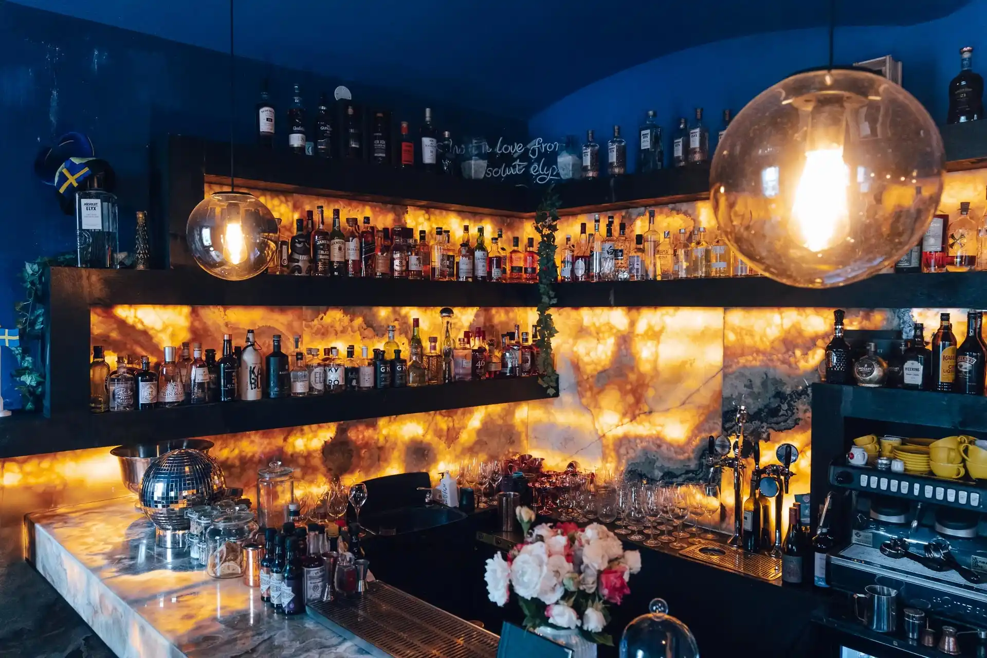 Eine stilvolle Bar mit leuchtenden, hinterleuchteten Regalen, auf denen verschiedene Schnapsflaschen, Gläser und Dekorationsartikel stehen. Große runde Leuchten hängen über dem Kopf, und ein Blumenstrauß steht auf dem Marmortresen im Vordergrund.