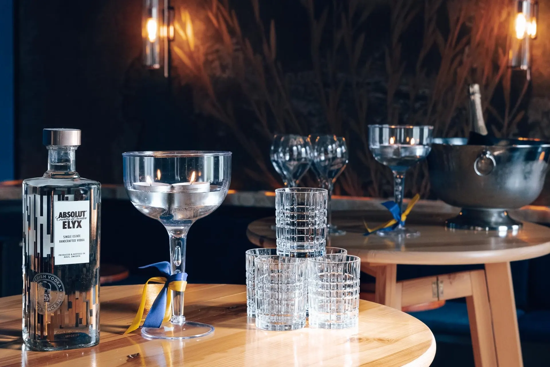 Eine Flasche Absolut Elyx Wodka, ein leeres Cocktailglas mit einer blauen und gelben Schleife und vier gestapelte Becher stehen auf einem Holztisch in einer schwach beleuchteten Bar mit weiteren Gläsern und einem Eiskübel im Hintergrund.