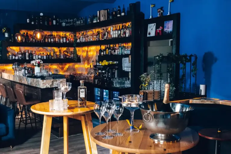 HEMMA Eine stilvolle Bar mit warmer Beleuchtung, Regalen mit Spirituosenflaschen und Gläsern sowie einem kleinen runden Tisch im Vordergrund mit einem Sektkübel, Weingläsern und einer Flasche Gin. Die Atmosphäre ist modern und einladend.