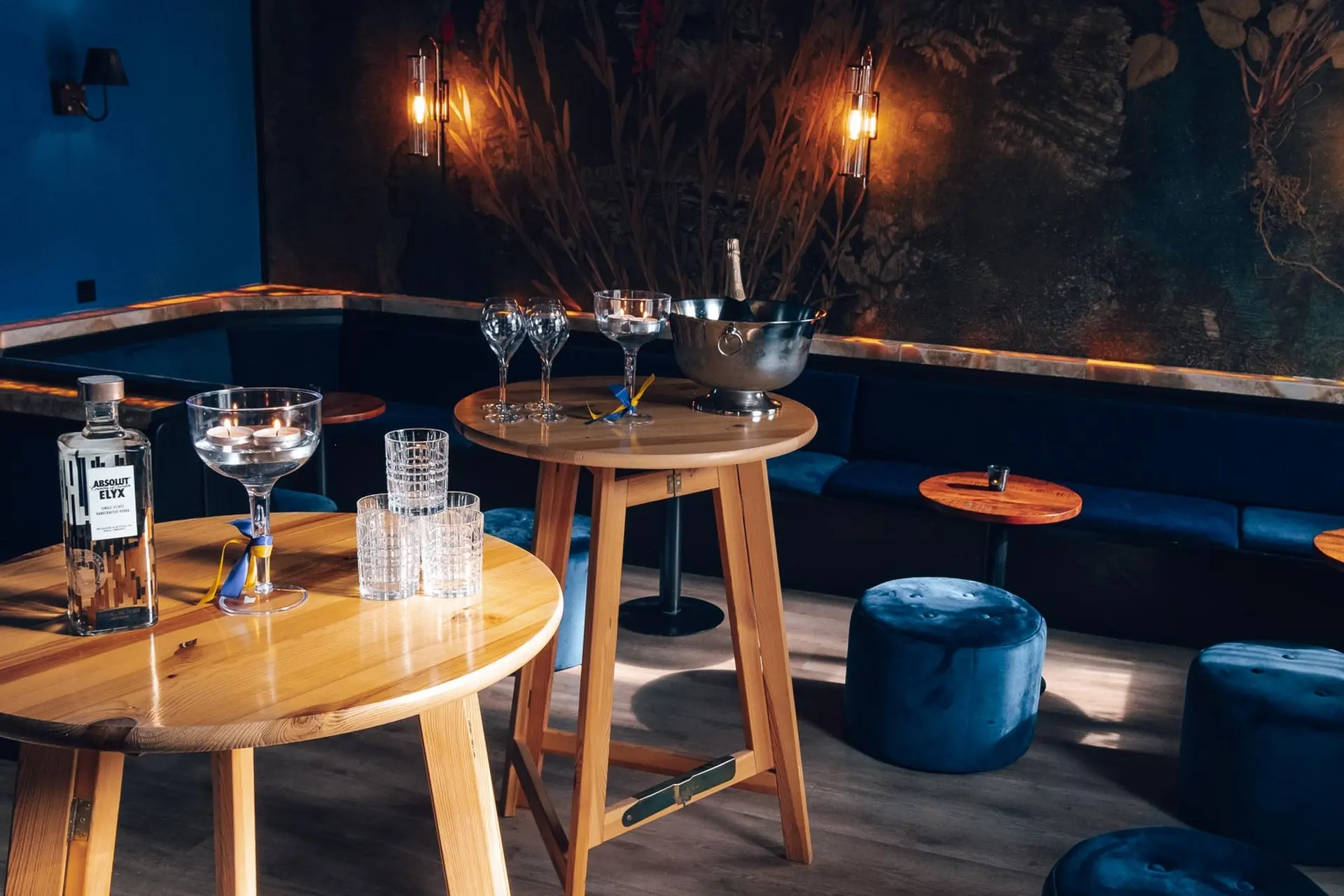 Eine stilvolle Lounge-Bar mit blauen Samtsesseln, runden Holztischen, Cocktailgläsern, einer Flasche Gin, einem Eiskübel und gedämpfter Wandbeleuchtung, die eine gemütliche, moderne Atmosphäre schafft.