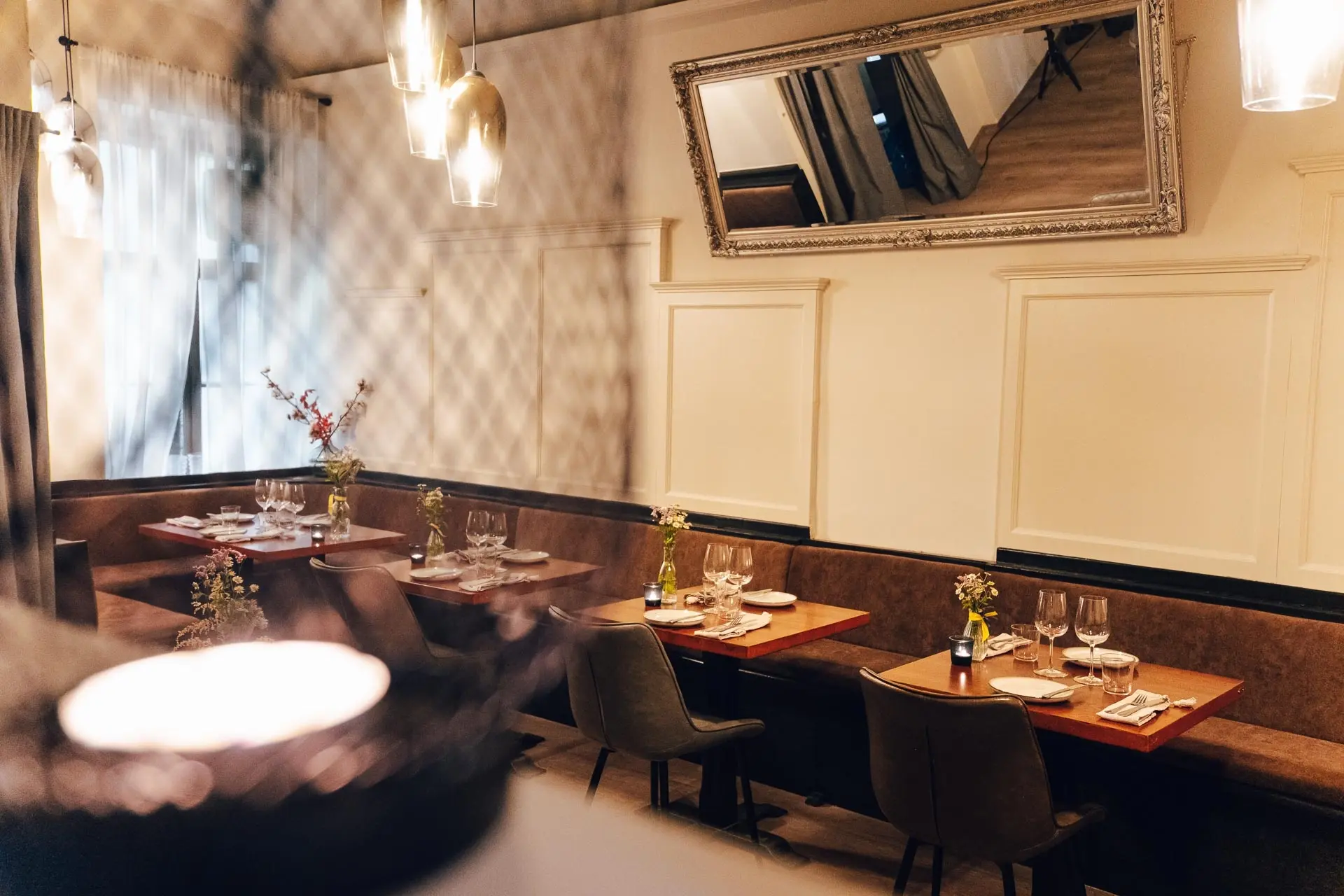 Ein gemütlicher Restaurantbereich mit braunen gepolsterten Bänken, gedeckten Tischen mit Gläsern und Blumen, warmer Hängebeleuchtung und einem großen Spiegel an der Wand über den Sitzplätzen.