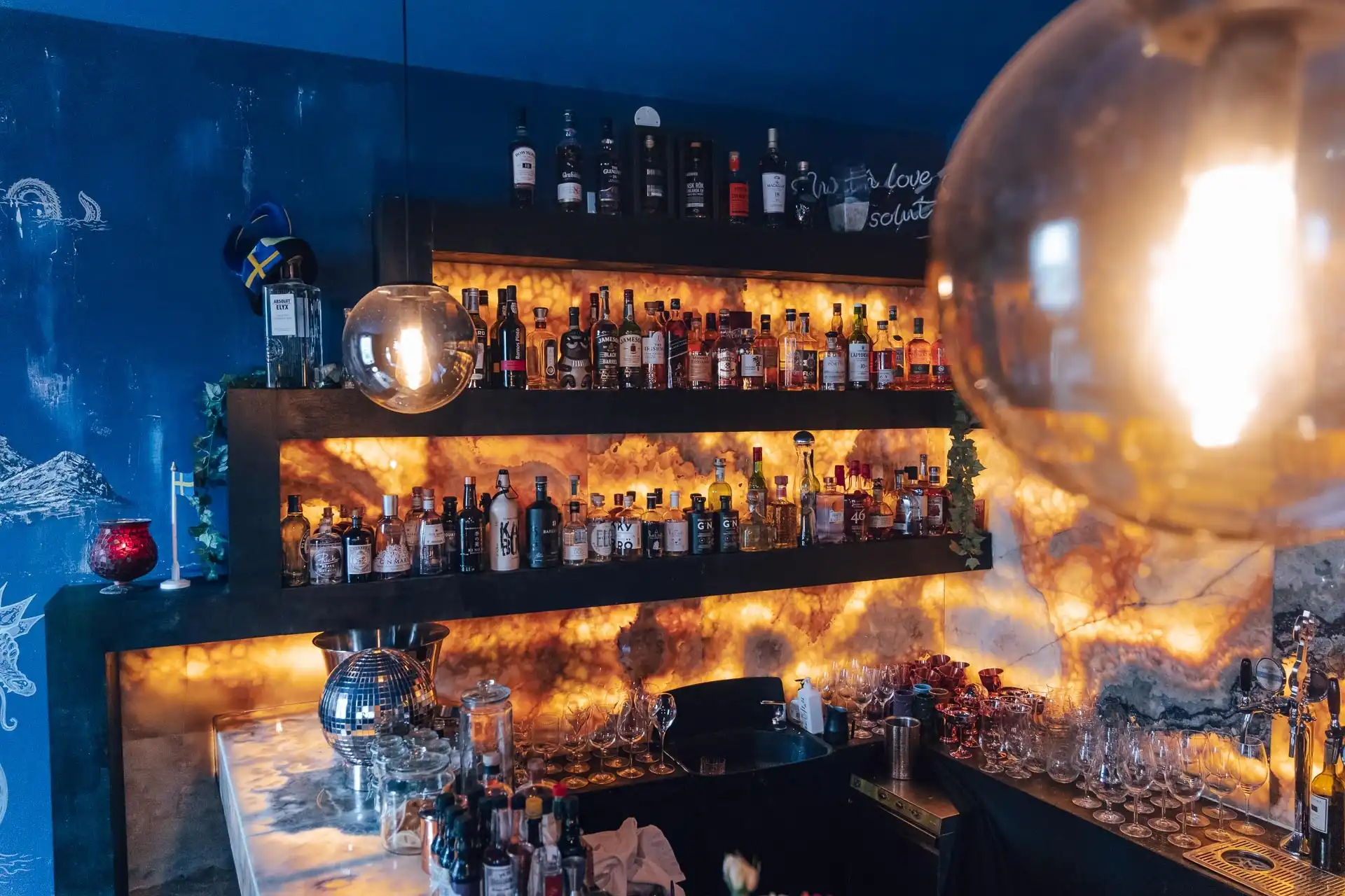 Eine stilvolle Bar mit beleuchteten Regalen mit verschiedenen Alkoholflaschen, Gläsern auf dem Tresen, hängenden Kugellampen und einer blauen Wand mit künstlerischen Zeichnungen im Hintergrund.