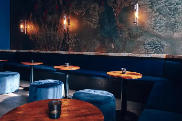 Eine stilvolle Lounge mit runden Holztischen, blauen Samthockern, einer langen blauen gepolsterten Bank, Wandleuchten und einem dekorativen Wandgemälde mit Blättern im Hintergrund. Jeder Tisch hat einen kleinen Kerzenständer.