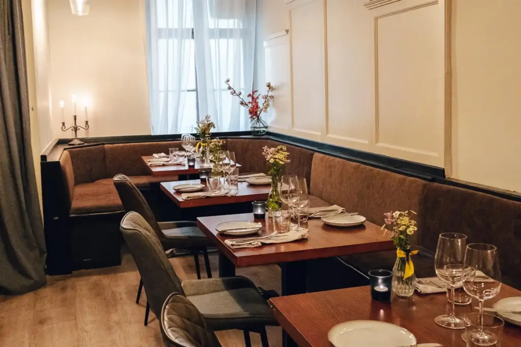 Ein gemütlicher Restaurant-Essbereich mit braunen gepolsterten Sitzbänken, Holztischen mit Tellern, Gläsern und Besteck, frischen Blumen in Vasen und sanftem natürlichem Licht aus einem Fenster mit durchsichtigen Vorhängen.