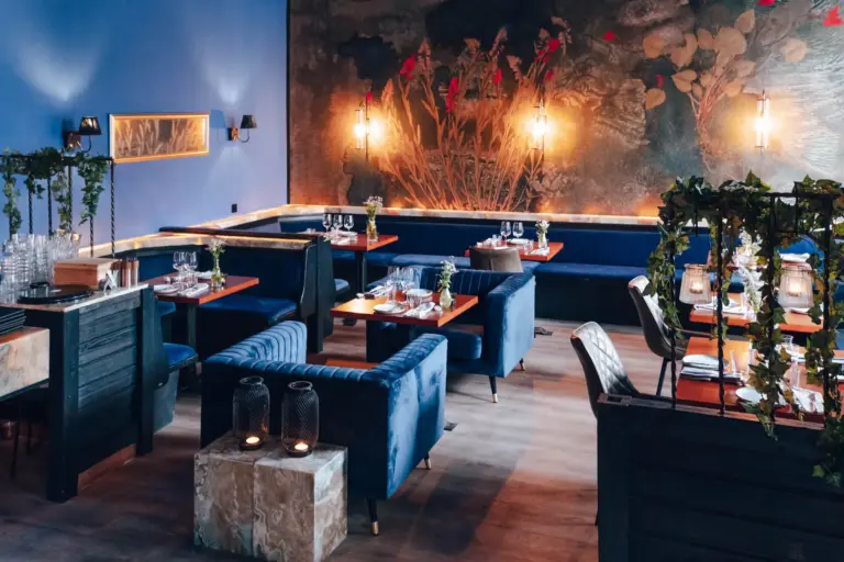 Hemma Ein stilvolles Restaurantinterieur mit blauen Samtstühlen, Holztischen zum Essen, Wandleuchten und einem großen Blumenwandbild an einer dunklen Wand. Grüne Pflanzen schmücken den Raum und schaffen ein gemütliches, elegantes Ambiente.