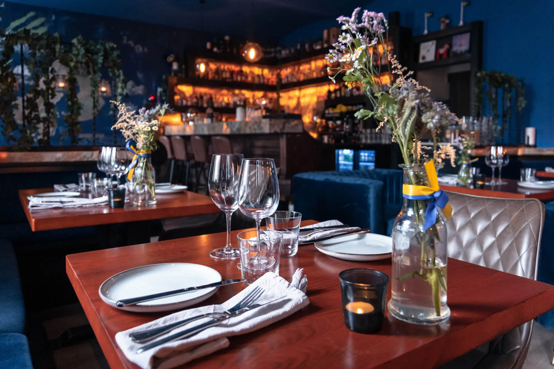 Ein gemütliches Restaurant mit Holztischen, die mit Tellern, Gläsern und Servietten gedeckt sind. Vasen mit Blumen und brennende Kerzen schmücken die Tische. Die Bar im Hintergrund erstrahlt in warmem Licht.