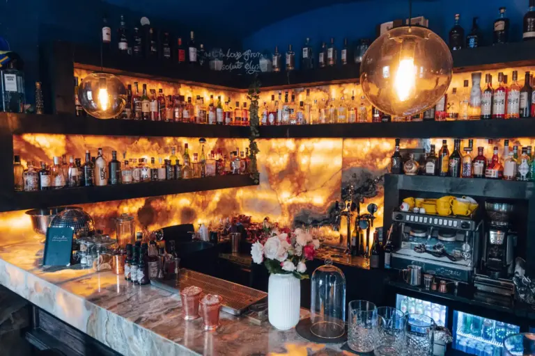 Eine stilvolle Bar mit leuchtenden, hintergrundbeleuchteten Marmorregalen, auf denen verschiedene Spirituosenflaschen stehen, eine Marmorarbeitsplatte mit Gläsern, eine Blumenvase und warme Pendelleuchten schaffen eine gemütliche Atmosphäre.