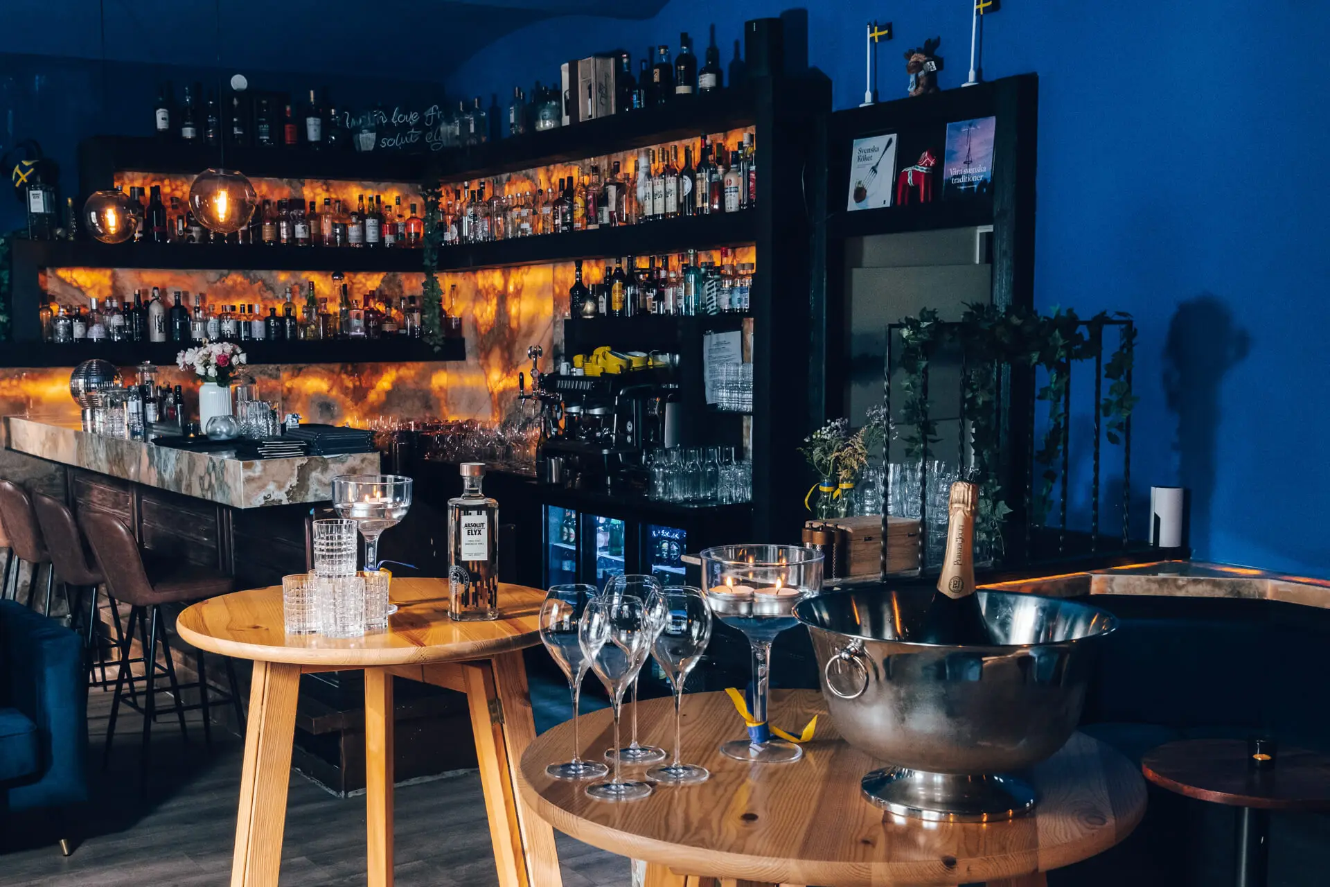 Eine stilvolle Bar mit Regalen voller Schnapsflaschen, orangefarbener Beleuchtung, einem Marmortresen und zwei runden Holztischen mit Gläsern, einer Champagnerflasche in einem Eiskübel und leeren Weingläsern.