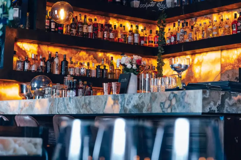 Eine stilvolle Bar mit beleuchteten Regalen mit verschiedenen Spirituosenflaschen, Gläsern und einer Marmorarbeitsplatte. Das Ambiente ist warm mit goldener Beleuchtung und elegantem Dekor, darunter Blumen und eine Discokugel.