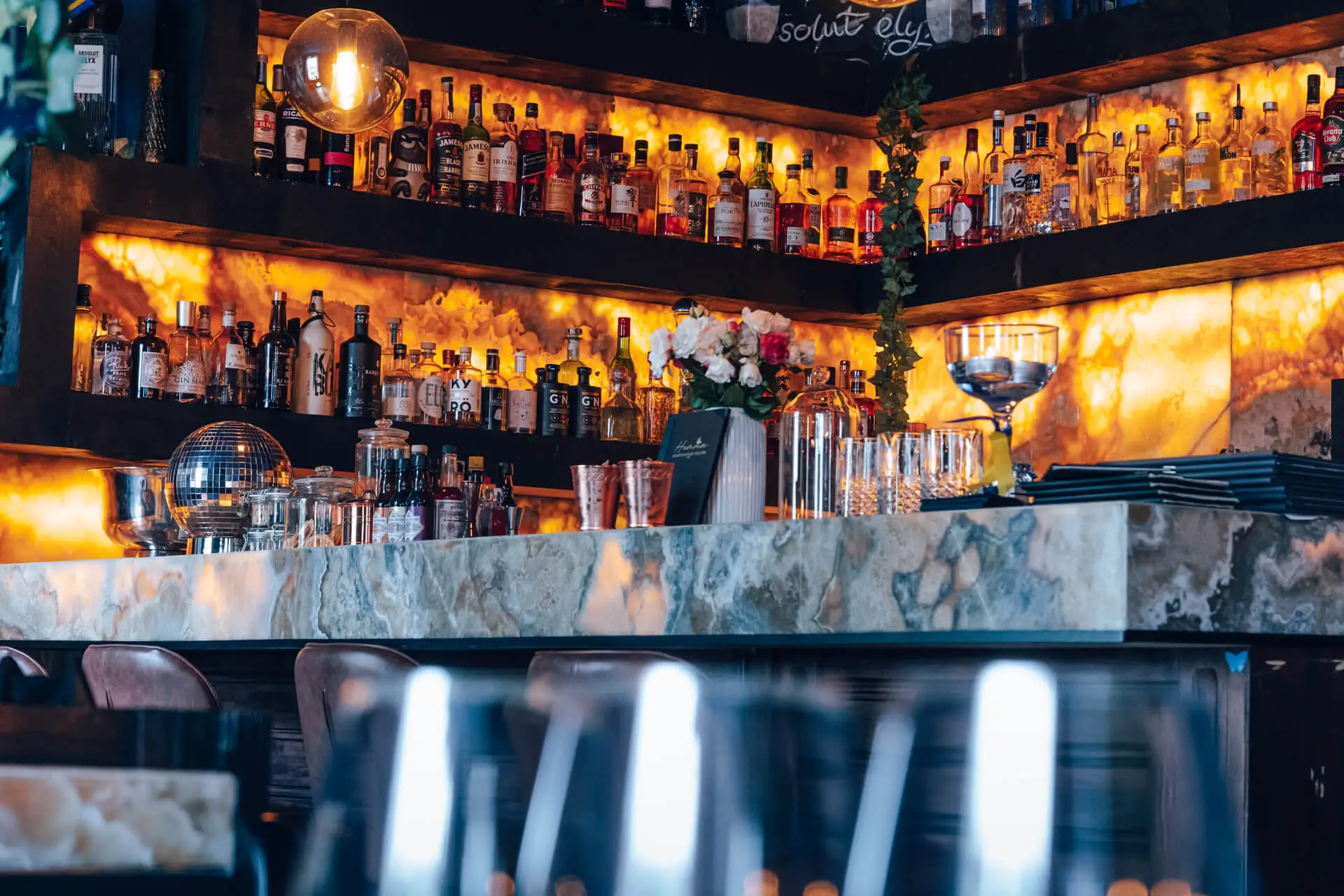 Eine stilvolle Bar mit beleuchteten Regalen mit verschiedenen Spirituosenflaschen, Gläsern und einer Marmorarbeitsplatte. Das Ambiente ist warm mit goldener Beleuchtung und elegantem Dekor, darunter Blumen und eine Discokugel.