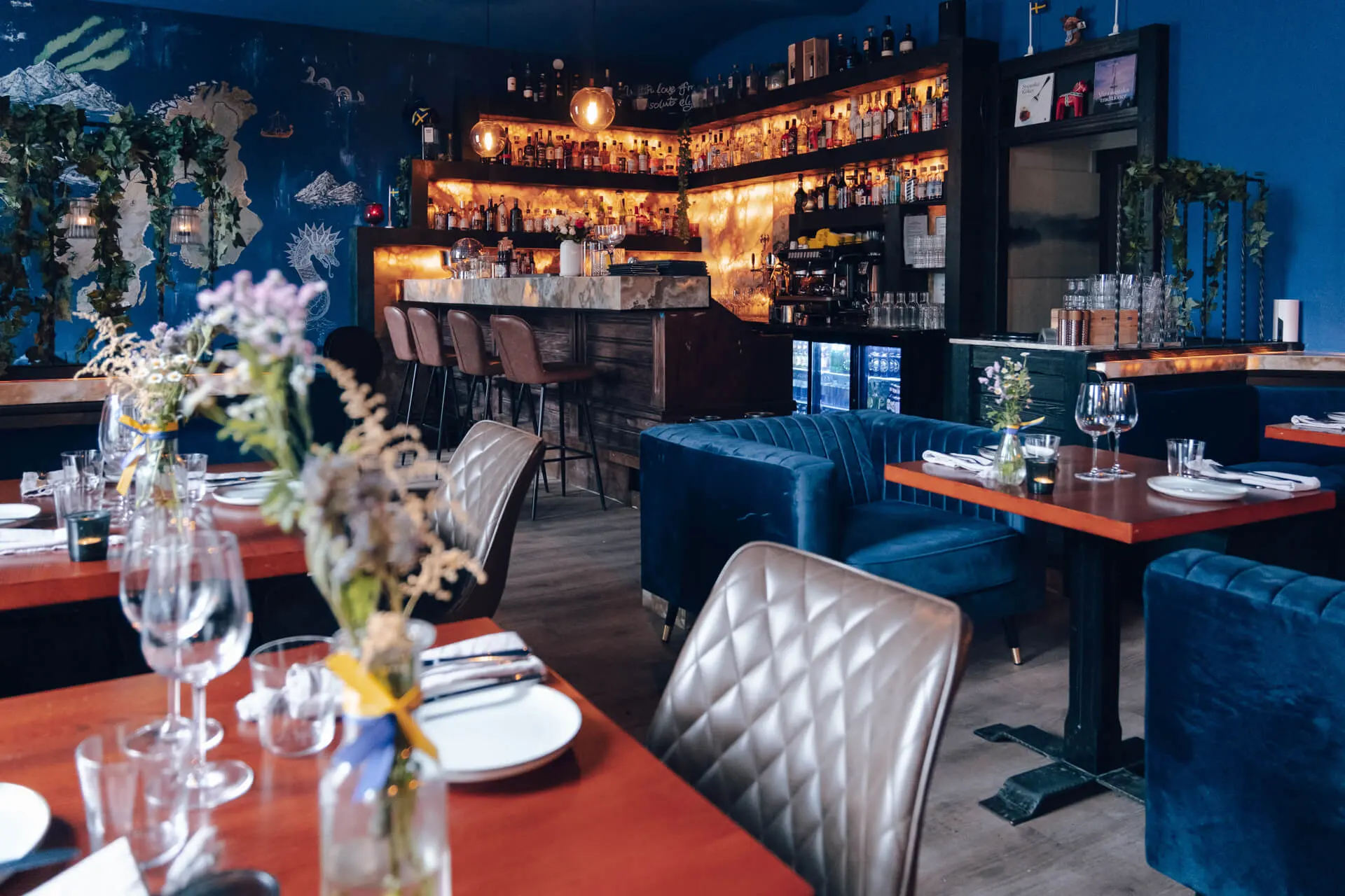 Ein gemütliches, stilvolles Restaurant mit blauen Wänden und Samtstühlen, Holztischen mit Tellern und Gläsern und einer gut beleuchteten Bar mit Flaschen und Hängelampen im Hintergrund.