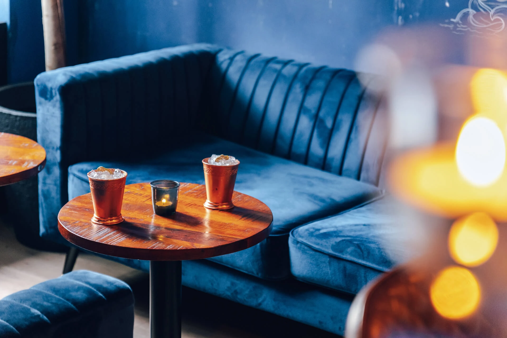 Eine gemütliche Lounge mit einem plüschigen blauen Samtsofa, einem Holztisch mit zwei Getränken in Kupferbechern mit Eis und einer brennenden Kerze, vor einer tiefblauen Wand.