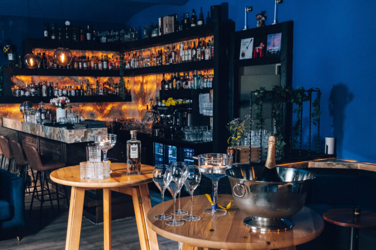 Eine stilvolle Bar mit Regalen voller Schnapsflaschen, einem Marmortresen, Hängelampen und einem runden Holztisch mit Weingläsern, einer Champagnerflasche in einem Eiskübel und Kerzen vor einer blauen Wand.
