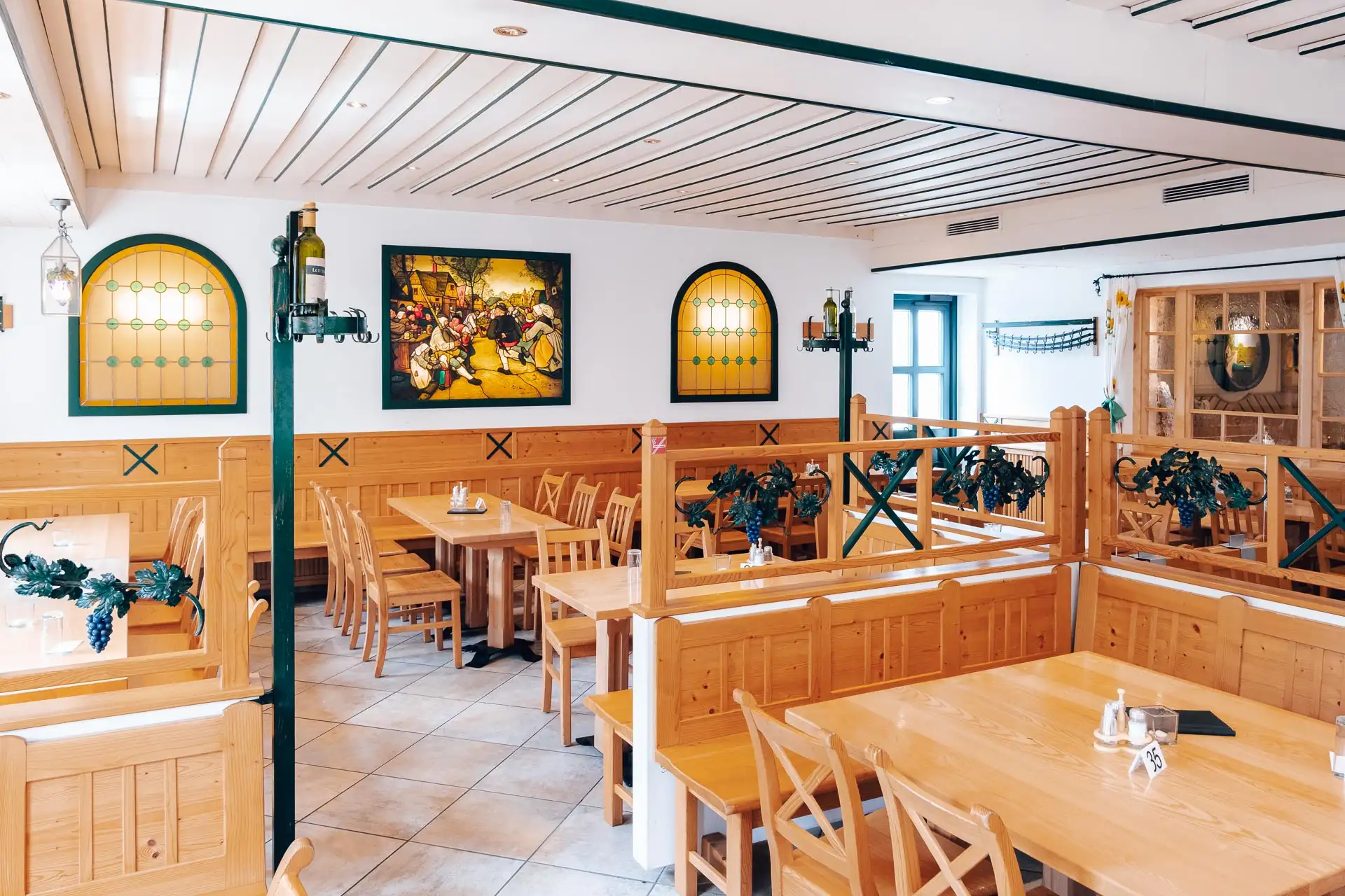 Ein helles, sauberes Restaurant mit hellen Holztischen und -stühlen, dekorativen Pflanzen, Wandkunst und gewölbten Buntglasfenstern. Die Tische sind einfach gedeckt, und der Raum hat eine gemütliche, einladende Atmosphäre.