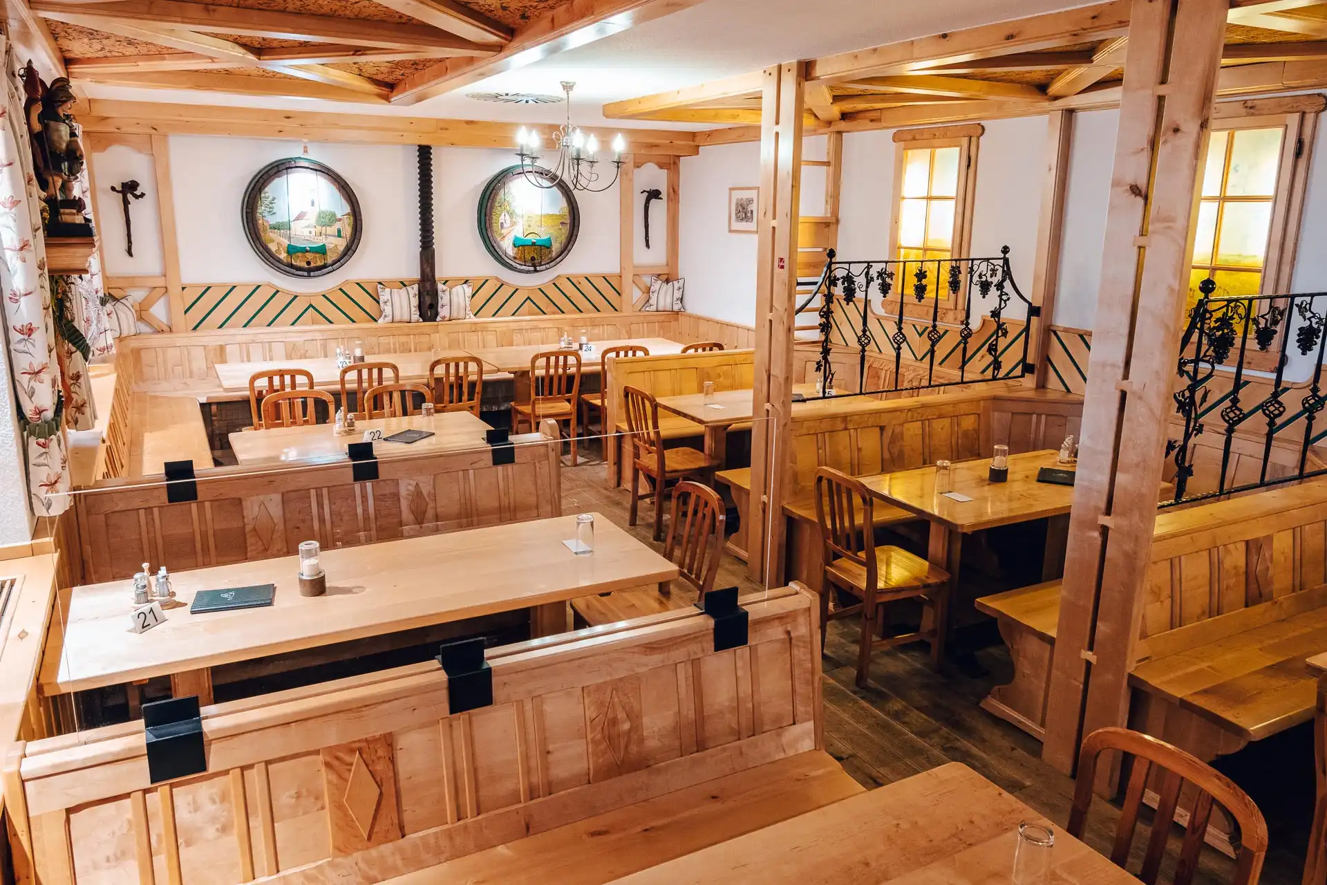 Ein gemütliches Restaurant mit Holztischen, -bänken und -stühlen. Holzbalken, gerahmte Spiegel an weißen Wänden, sanfte Beleuchtung und dekorative Eisengeländer schaffen eine warme, rustikale Atmosphäre.