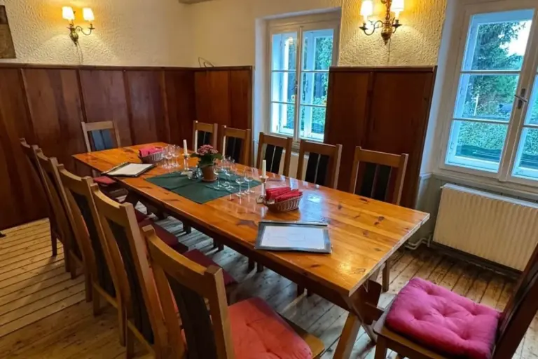 Ein hölzerner Esstisch mit acht Stühlen, einige davon mit roten Kissen, in einem gemütlichen Raum mit holzgetäfelten Wänden, einem Fenster und Wandleuchten, die zum Essen aufgestellt sind. Auf dem Tisch stehen Gläser, Speisekarten und eine Topfpflanze.