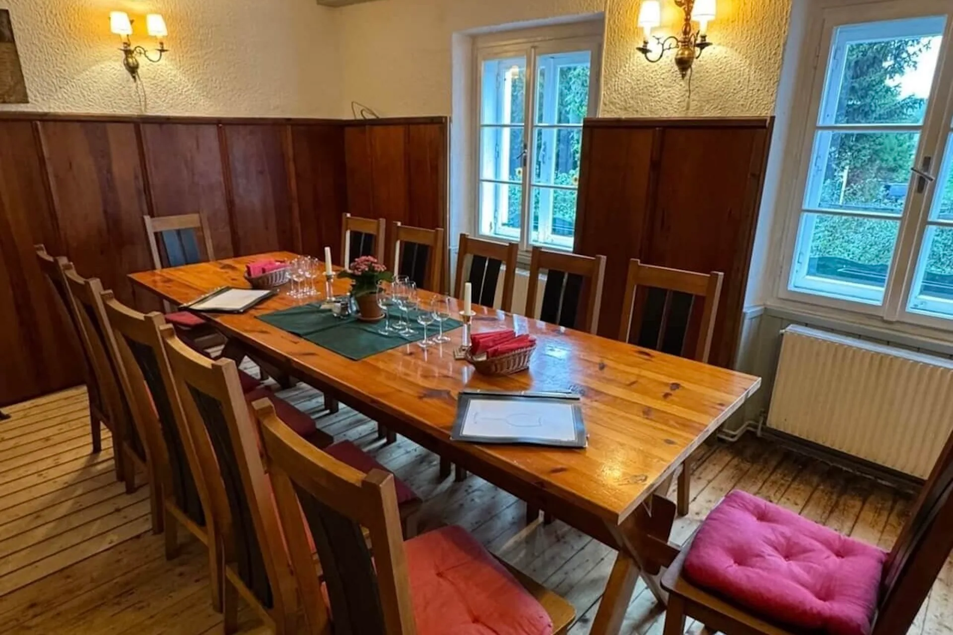 Ein hölzerner Esstisch mit acht Stühlen, einige davon mit roten Kissen, in einem gemütlichen Raum mit holzgetäfelten Wänden, einem Fenster und Wandleuchten, die zum Essen aufgestellt sind. Auf dem Tisch stehen Gläser, Speisekarten und eine Topfpflanze.