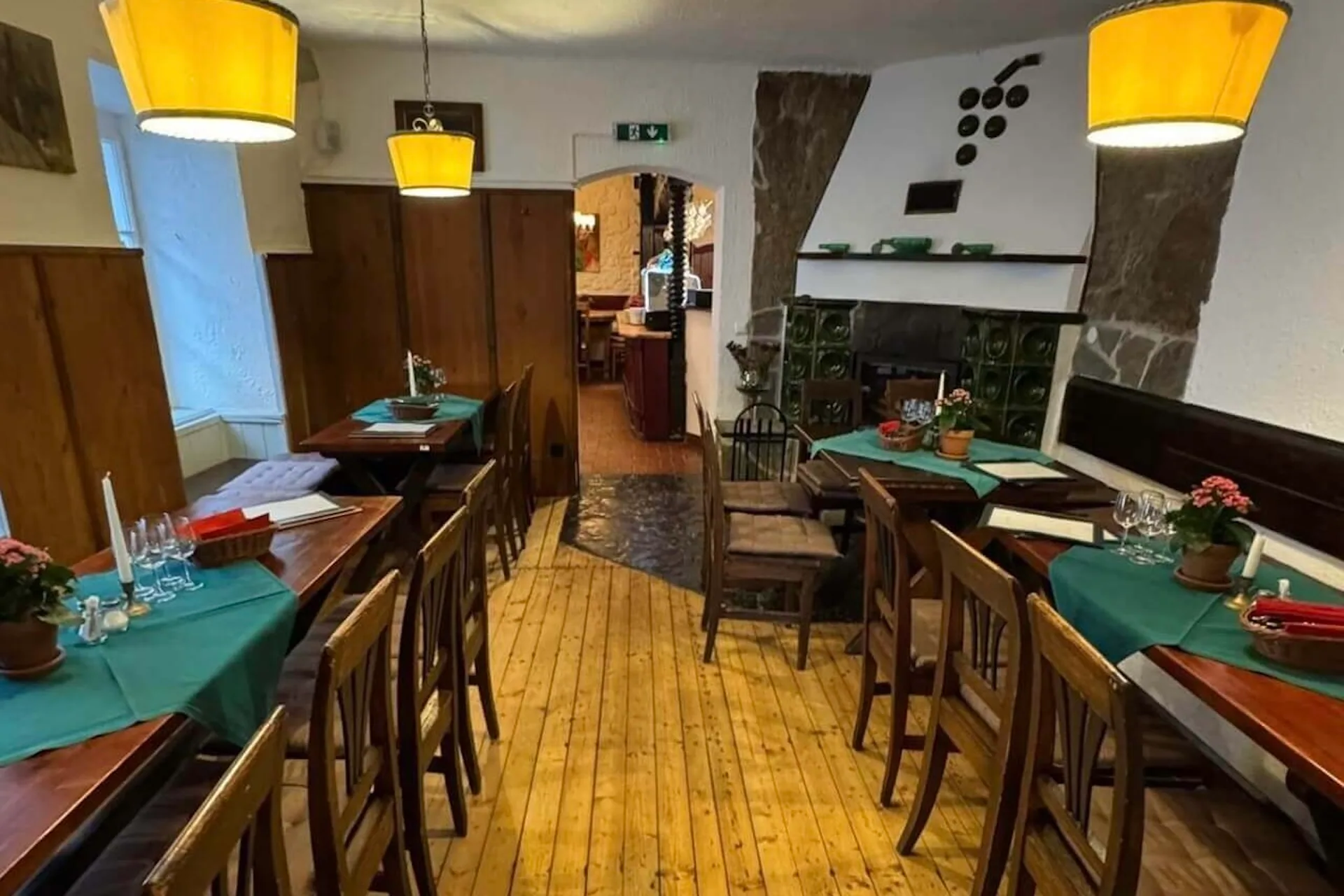 Ein gemütliches Restaurant mit Holztischen und -stühlen, grünen Tischsets, sanfter gelber Beleuchtung und einem Kachelofen an der Wand. Im gesamten Essbereich sind Pflanzen und Tischgedecke fein säuberlich angeordnet.