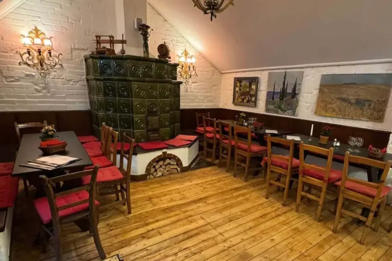 Gemütliches Restaurantinterieur mit Holzboden, Tischen und Stühlen mit roten Kissen, einem grünen Kachelofen in der Ecke, dekorativen Lampen und Gemälden auf weißem Putz und holzgetäfelten Wänden.