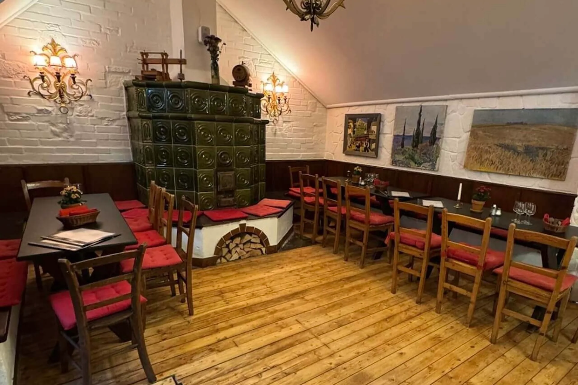 Gemütliches Restaurantinterieur mit Holzboden, Tischen und Stühlen mit roten Kissen, einem grünen Kachelofen in der Ecke, dekorativen Lampen und Gemälden auf weißem Putz und holzgetäfelten Wänden.